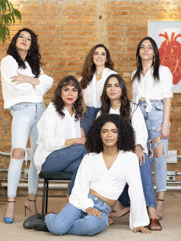 Foto de seis mulheres. Uma a frente no chão, duas atrás sentadas, e três em último plano de pé. Todas estão de camisa branca e jeans