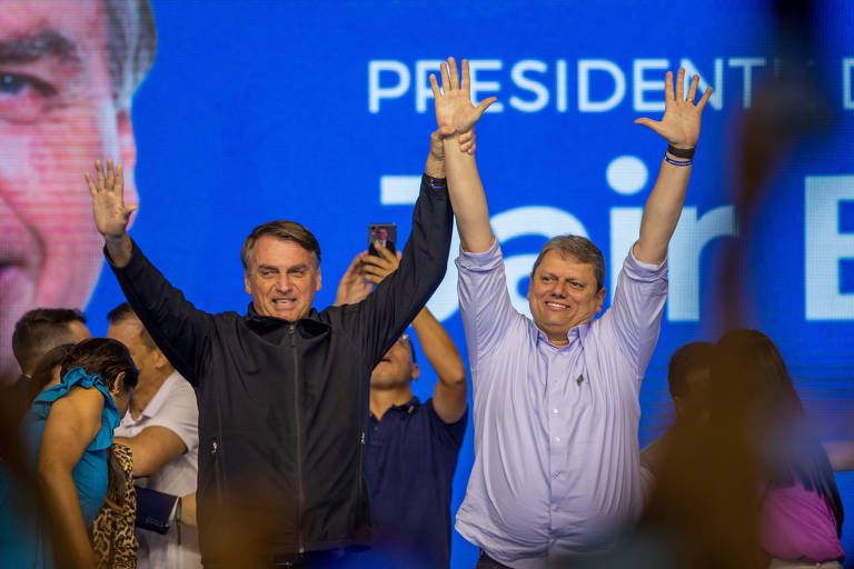 Diante de uma tela com fundo azul, com os dizeres Jair Bolsonaro e uma foto do presidente, Bolsonaro e Tarcísio, dois homens brancos, cabelos castanhos, estão lado a lado com os braços erguidos. Há várias pessoas ao fundo