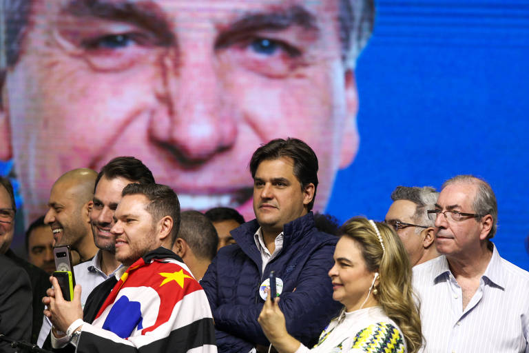 Várias pessoas aglomeradas em um palco que tem ao fundo a foto de Bolsonaro. Entre elas, Silveira, um homem branco, jovem e careca, que sorri, e Cunha, no canto direito, um homem branco, calvo, cabelos brancos, óculos e camisa branca com a gola aberta