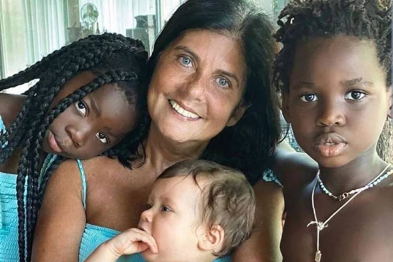 Senhora branca sorrindo com bebê branco no colo, uma menina preta a sua esquerda e um menino preto a direita
