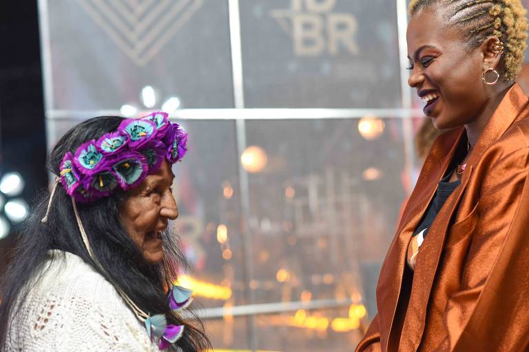 Indígena idosa com flores na cabeça sorri ao lado de mulher negra alta com jaqueta laranja