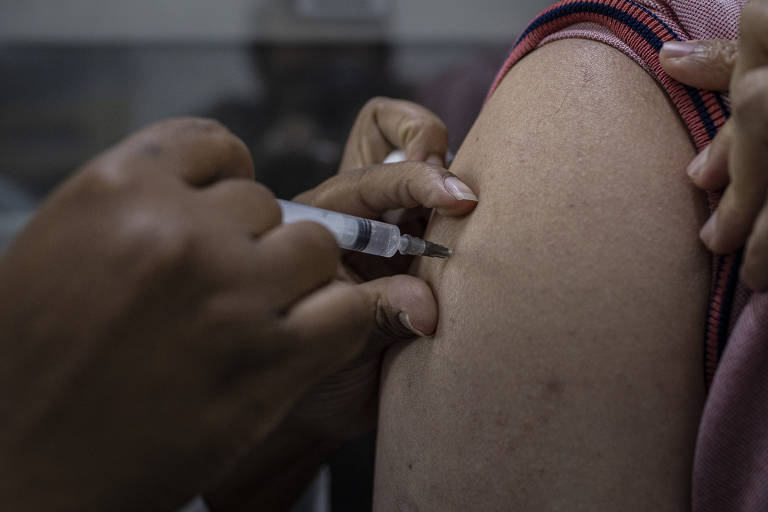 Mãos de pessoa negra vacinam braço de pessoa branca. Foto está bem fechada nesta ação