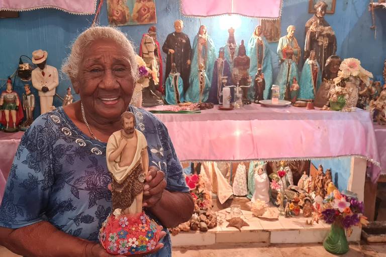 Mulher negra mais velha segura imagem de São João rodeada por altar cheia de santos