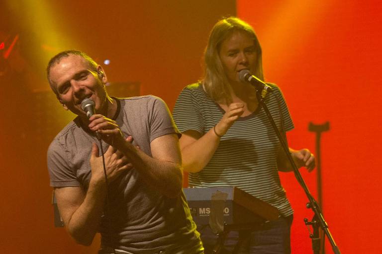 Stuart Murdoch, vocalista da banda escocesa Belle and Sebastian, está no palco com camiseta e calça cantando com um microfone