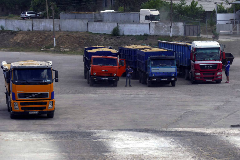 Imagem mostra quatro caminhões parados em um pátio. Os caminhões são das cores amarelo, laranja, azul e vermelho. Há dois homens parados em pé perto dos caminhões.