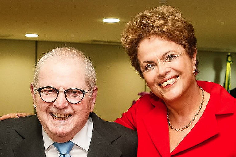 Jô Soares, de terno azul escuro e gravata azul clara, aparece ao lado de ex-presidente Dilma Rousseff, de vermelho; ela passou o braço sobre os ombros dele