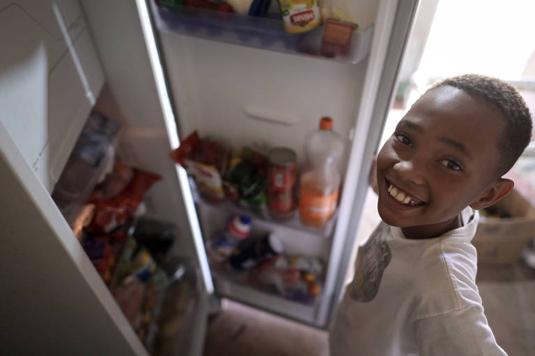 O menino segura a porta da geladeira aberta e mostra que ela está cheia de alimentos