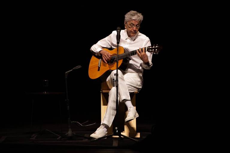 Caetano Veloso usa roupa branca e toca um violão num palco