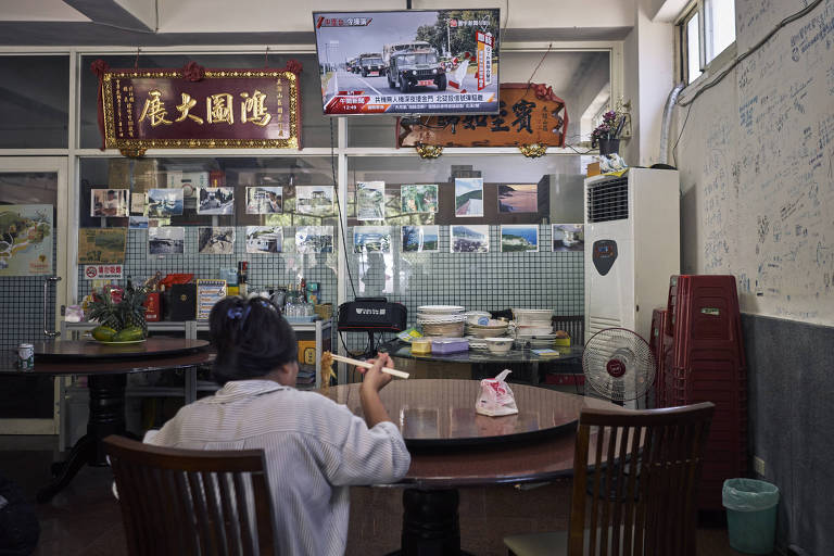 Fotografia colorida mostra mulher sentada de costas para a câmera à mesa de um restaurante; ela veste camisa branca e tem os cabelos pretos e lisos presos, enquanto segura um hashi; na TV à sua frente, no alto, noticiário sobre exercícios militares chineses