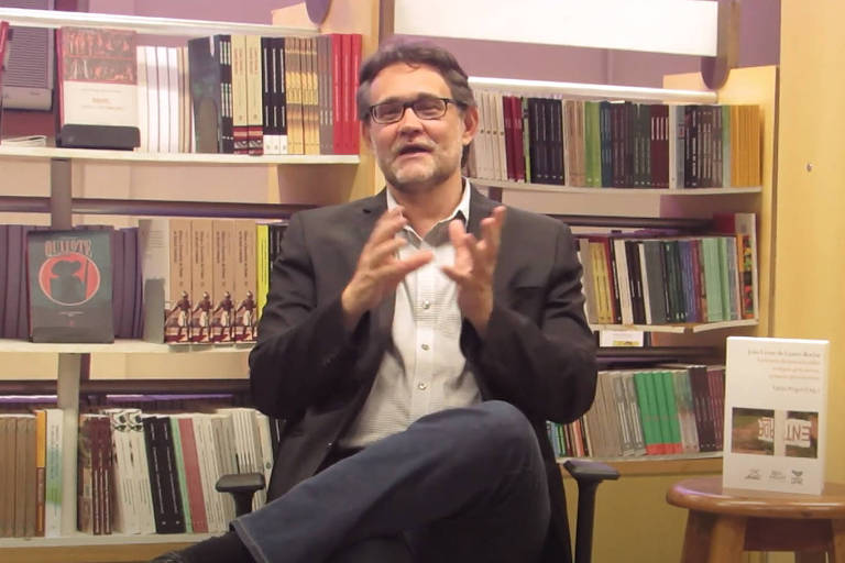 O professor João Cezar de Castro Rocha sentado em frente a uma estante cheia de livros