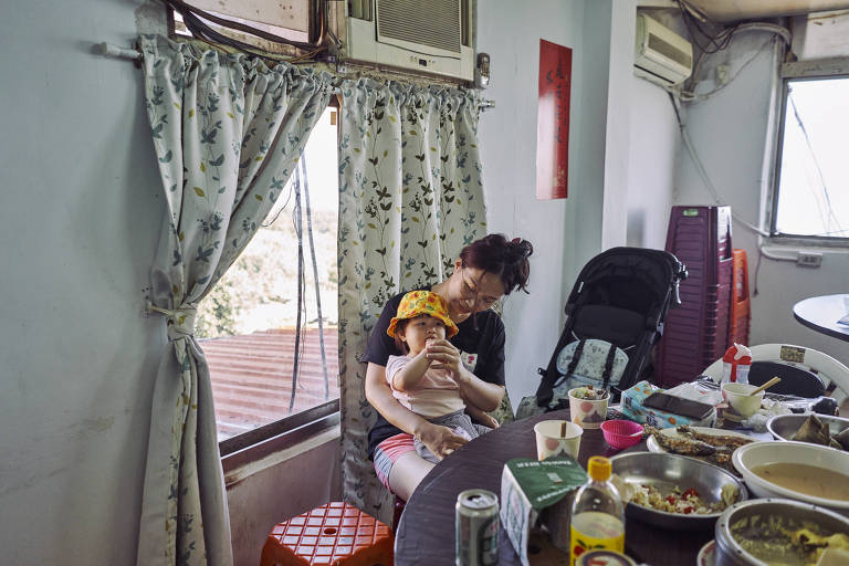 Fotografia colorida de mulher asiática de origem taiwanesa com filha de dois anos de idade no colo; elas estão sentadas a uma mesa cheia de pratos e recipientes de comida