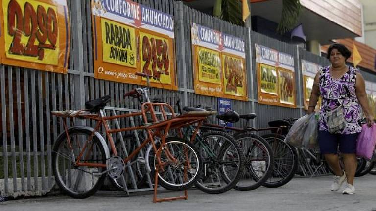 Fotografia colorida mostra uma calçada de um supermecado; em primeiro plano estão bicletas estacionadas com anúncio de promoções de um supermercado afixadas em uma grade; em segundo plano, caminha uma mulher negra com sacolas de compras nas mãos