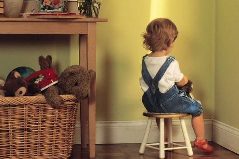 Criança está em um quarto, sentada de frente para uma parede