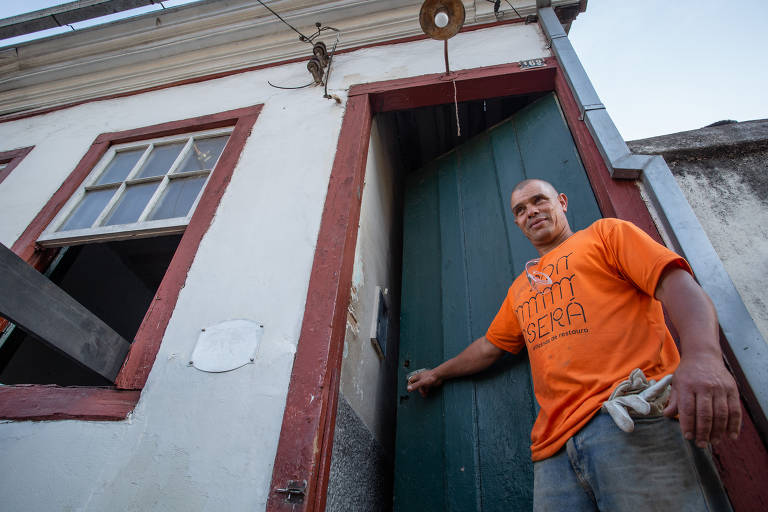Homem de camiseta laranja, luvas apoiadas em calça jeans, abre a porta azul de casa histórica com detalhes em vermelho na janela e na porta