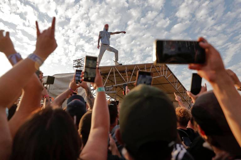 Imagem mostra cantor em cima de uma estrutura de metal. Embaixo, há vários fãs com os celulares apontados para cima.