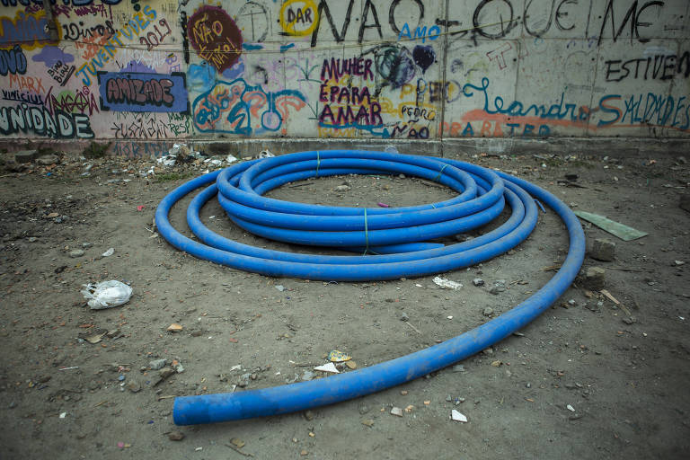 Imagem mostra cano azul em um chão de terra. Ao fundo, há um muro cheio de grafite e pichações.