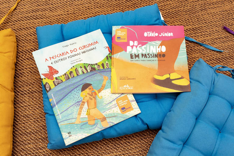 Dois livros infantis coloridos estão em cima de uma almofada azul; um dos livros tem um indigena na capa e o outro  o pé de uma menina