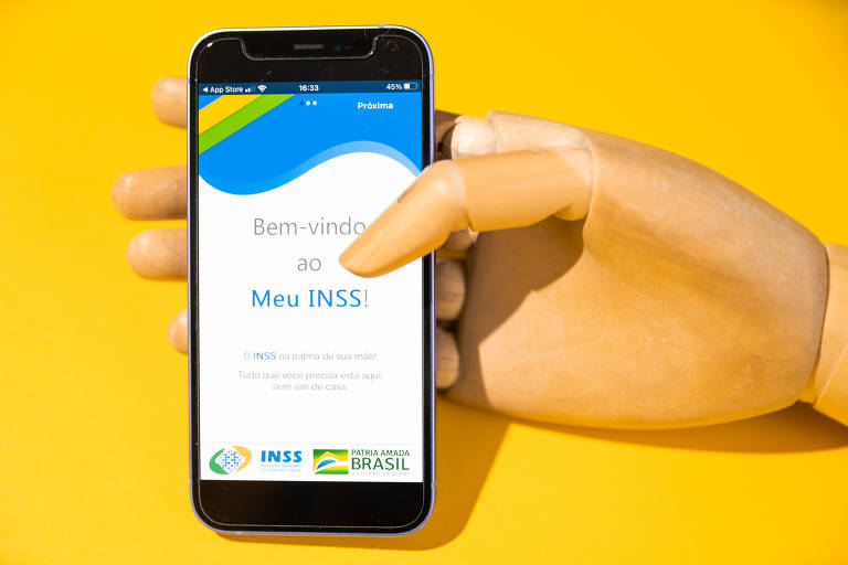 Tela de celular com o aplicativo Meu INSS aberto. Mão de madeira a segura, na frente de fundo amarelo