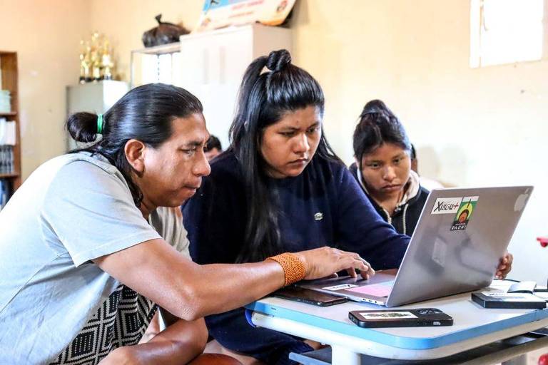 Duas jovens indígenas ao lado de um homem indígenas sentados em frente a um laptop participando de uma atividade on-line