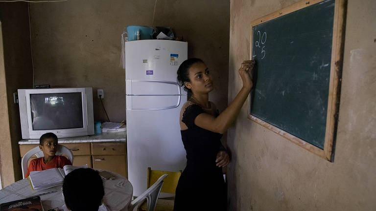 Foto colorida retrata um cômodo de uma casa. Há uma televisão, uma geladeira, uma mesa onde duas crianças estão sentadas com livros e cadernos abertos e uma lousa pendurada em uma parede, onde uma mulher de vestido preto escreve o abecedário com um giz.