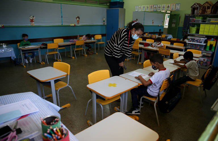 Professora orienta aluno sentado em carteira; há poucos estudantes em sala e todos usam máscara contra a Covid-19