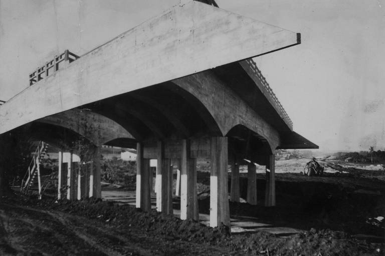 Foto em preto e branco da construção de uma rodovia. É possível ver uma estrutura de concreto com vários arcos dentro sendo construída. No chão, parece ser gramado