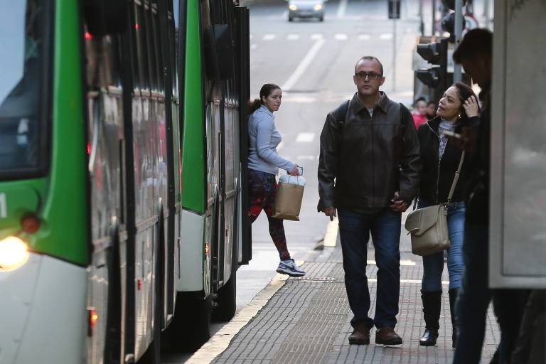 Imagem mostra casal caminhando de mãos dadas em uma calçada, em frente a dois ônibus verdes que estão parados. Há mais pessoas no ponto de ônibus.