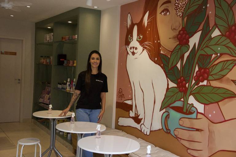 Mariana na cafeteria em frente a parede com desenho em que uma mulher está abraçada com um gato e um pé de café
