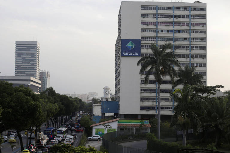 Imagem mostra foto de prédio da Estácio no Rio de Janeiro. Ele é branco e tem uma placa com a logo da universidade. Ao lado, há uma rua com carros e ônibus. Ao fundo, é possível ver outro prédio, que está distante.