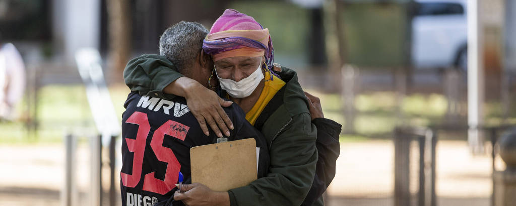 Mulher de máscara, casaco, lenço rosa na cabeça e prancheta na mão abraça homem com camisa de time, de costas