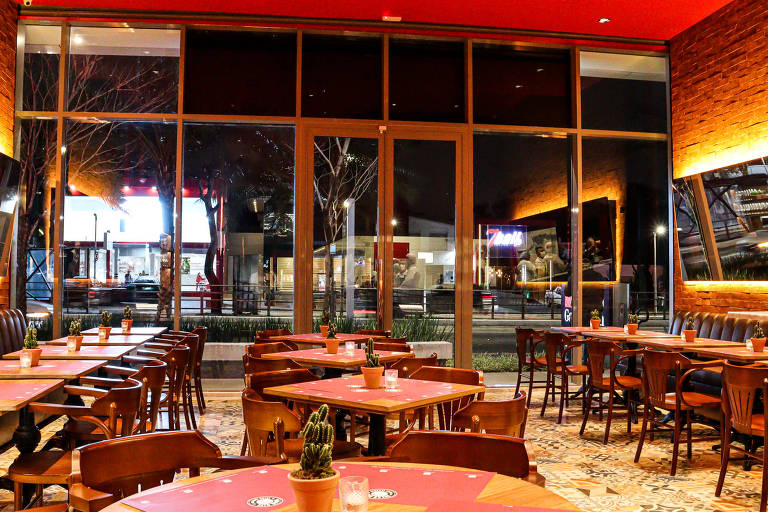 Salão de restaurante com paredes vermelhas, mesas de madeira, pé-direito alto e fachada envidraçada