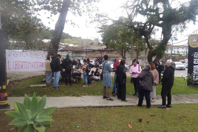 Grupo de pessoas aguarda em frente a uma delegacia, em um local arborizado. À esquerda, há uma faixa branca em que se lê escrito com spray preto e vermelho: "Abaixo à repressão" e "Libertem a estudante".