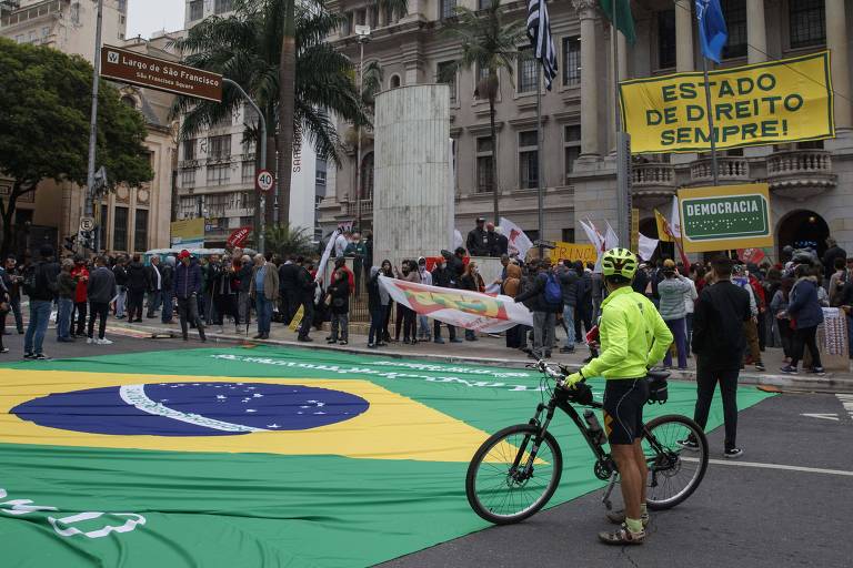 Homem e sua bicicleta em frente a uma bandeira do Brasil esticada no chão, em frente ao prédio da Faculdade de Direito