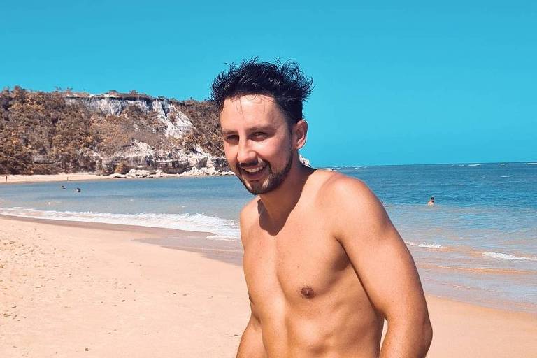 A imagem colorida mostra um homem branco, sem camisa em uma praia