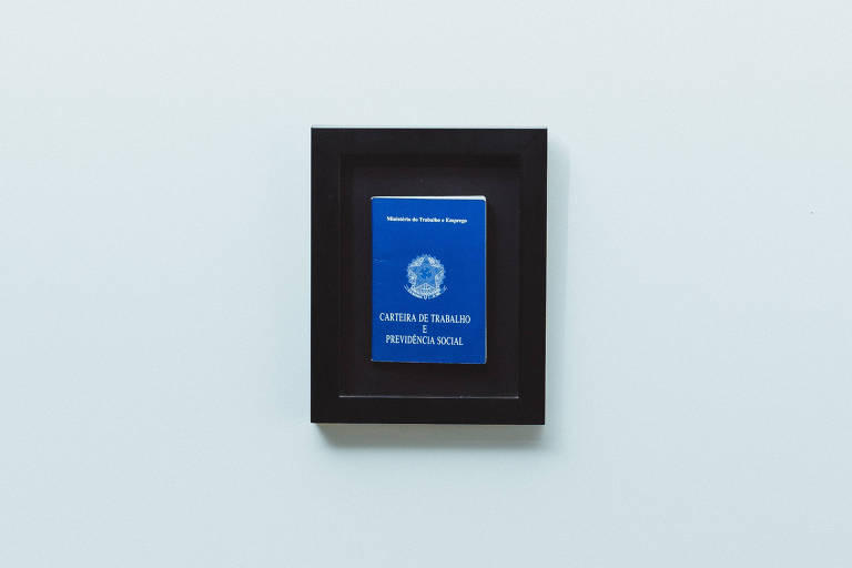 Foto mostra uma carteira de trabalho azul emoldurada num pequeno quadro preto sobre superficie azul clara