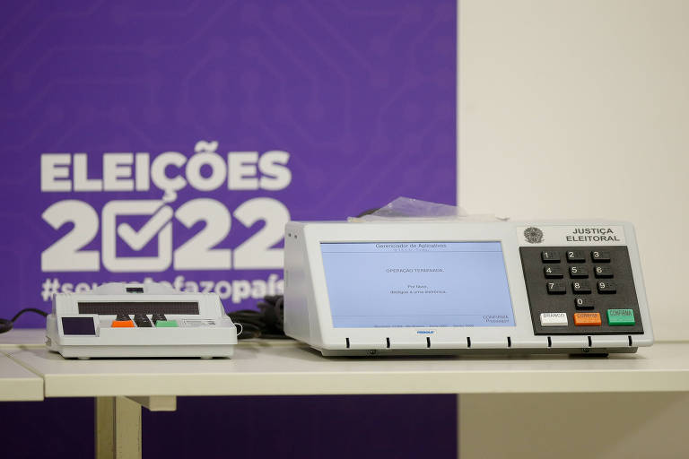 Sobre uma mesa há uma urna eletrônica. Ao fundo, se vê um cartaz escrito: Eleições 2022.