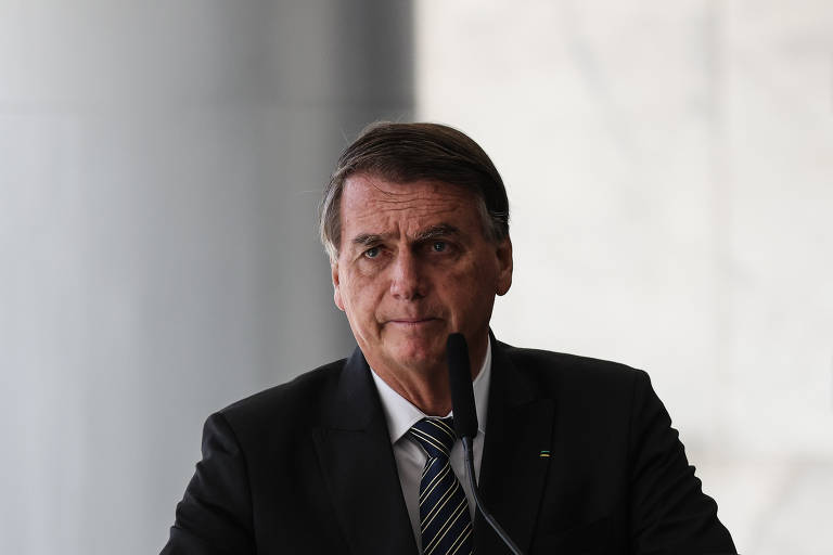 Imagem em primeiro plano mostra Jair Bolsonaro de roupa social, com semblante sério e na frente de um microfone
