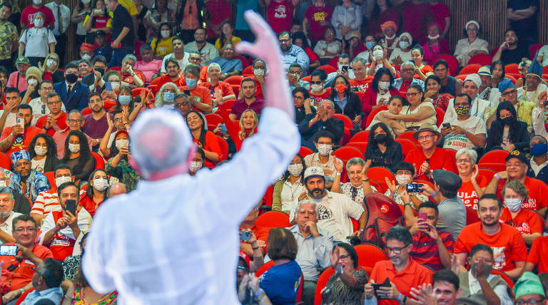 Lula, homem branco e com careca centralizada, está de costas, de camisa branca, com a mão direita levantada, com multidão ao fundo, de vermelho, em nível mais baixo