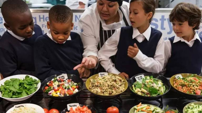Cozinheira explica para quatro crianças quais são os alimentos dispostos em pratos em uma mesa