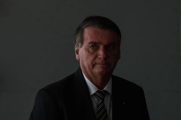 Em um fundo cinza, com uma sombra na parte esquerda do rosto, Bolsonaro, um homem branco, olhos claros, cabelos lisos e castanhos, vestido de terno, gravata e camisa branca