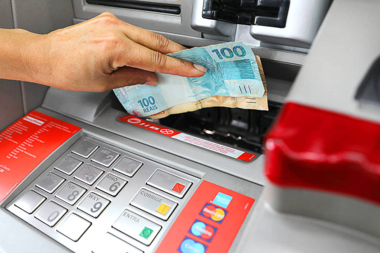Imagem aproximada mostra uma mão depositando cédulas de R$ 100 e R$ 50 em um caixa eletrônico do Banco24Horas.