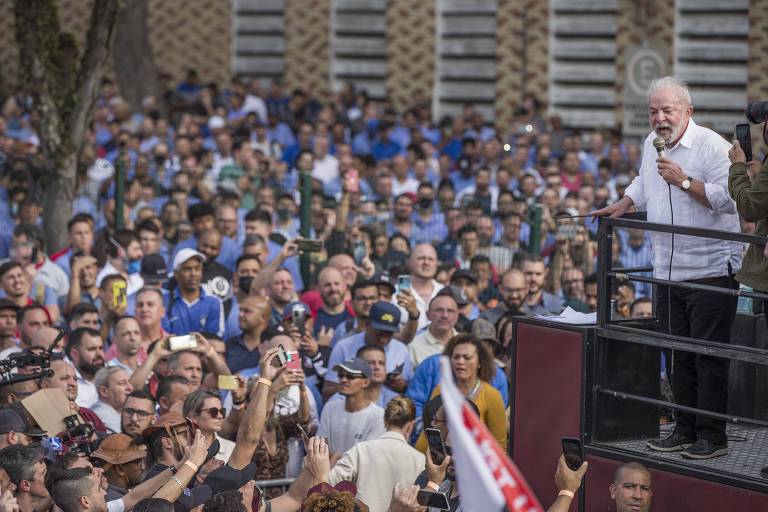Lula está em cima de um palanque e segura microfone junto à boca; dezenas de apoiadores se aglomeram e aparecem na imagem. Algumas pessoas seguram celulares enquanto observam o petista