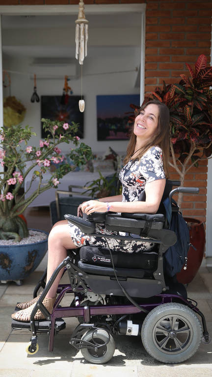 Mara Gabrilli está sentada em sua cadeira de rodas motorizada e sorri. Ela usa vestido florido e sandálias. Ao fundo, uma porta de varanda, vasos de flores e quadros