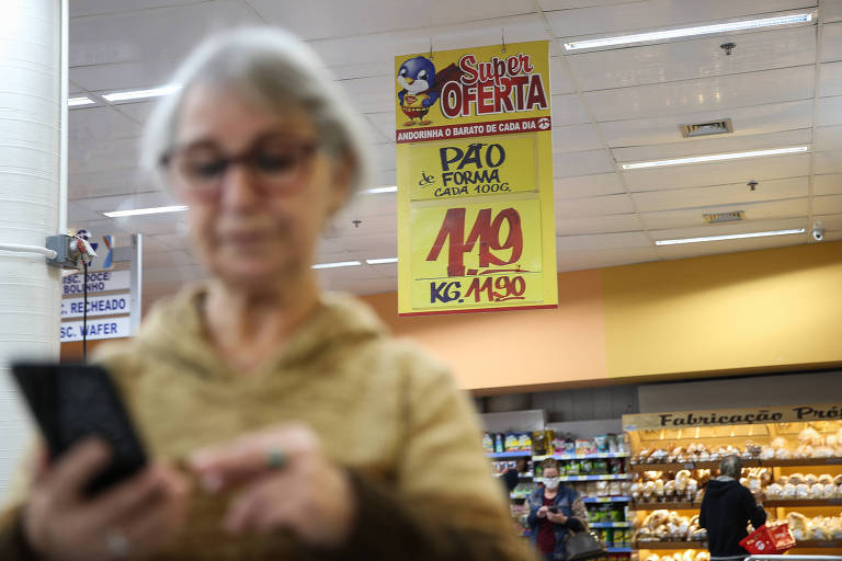Imagem mostra consumidora pesquisando no celular enquanto faz compras no supermercado
