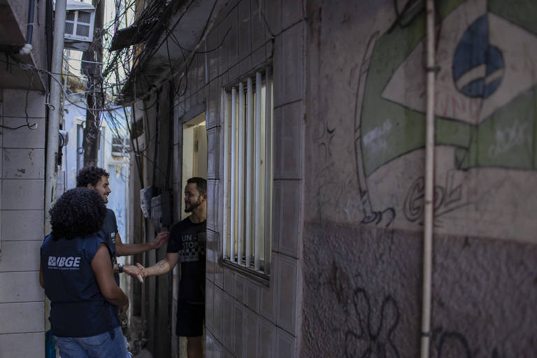 Dois recenseadores, um homem e uma mulher, usam colete do IBGE e cumprimentam morador na porta de uma casa com um aperto de mão