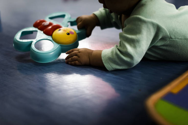 Imagem mostra bebê deitado de barriga para o chão segurando brinquedo em formato de borboleta.