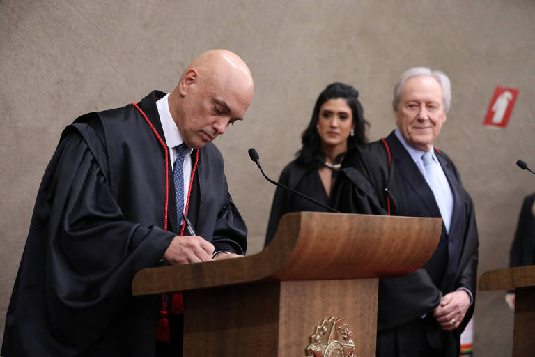 Moraes está curvado, sobre um púlpito, com uma caneta posicionada sobre papel; é observado por Ricardo Lewandowski, que sorri