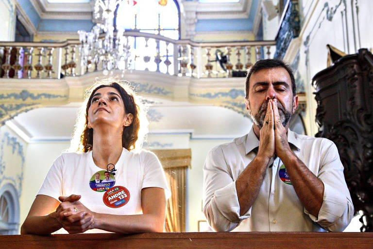 Imagem em primeiro plano mostra Marcelo Freixo com os olhos fechados e as mãos unidas na altura do rosto. Ao lado, está sua mulher, que olha para cima. Eles estão dentro de uma igreja.