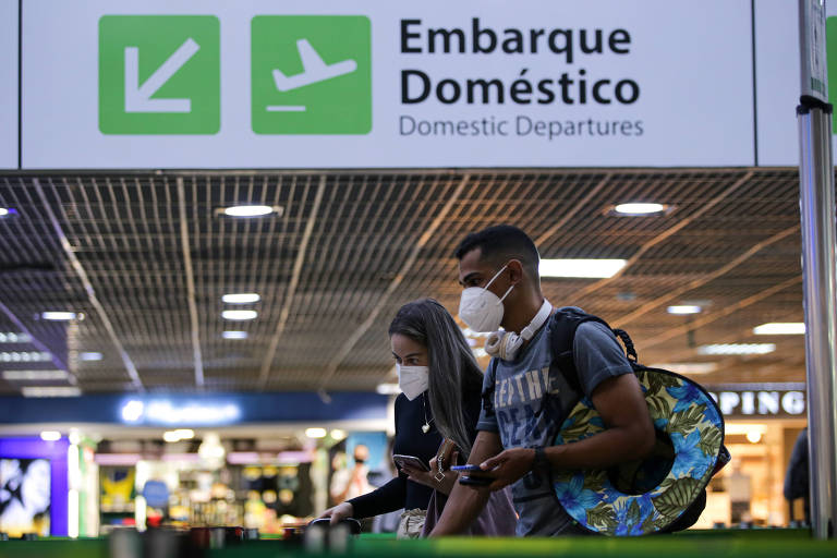 Mulher branca, loira e de cabelos lisos, caminhando ao lado de homem pardo, ambos de máscara no rosto e carregando malas em aeroporto. Acima dos dois, uma placa indicando o local de embarque doméstico.
