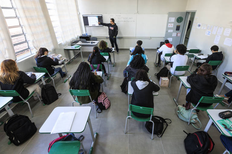 Alunos sentados em carteiras numa sala de aula, com uma professora explicando lição próximo à lousa